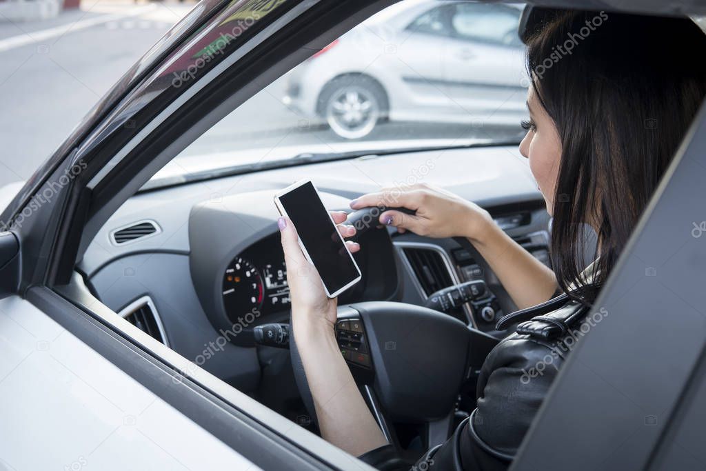 Side view of car in traffic with pretty female driver who has opened window and checks smartphone. Brunette girl holding mobile with black screen and driving. Risky driving. Distracted female driver.