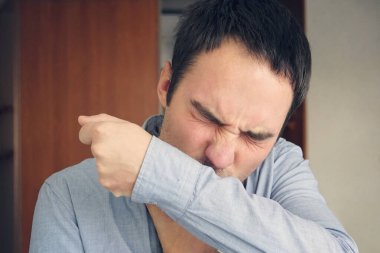 Adam grip olmuş ve kolunun kıvrımlarında koluna hapşırıyor. Genç bir adam viral bakterilerin yayılmasını önlemek ve diğer insanlara bulaştırmamak için gömleğinin dokusuna öksürüyor. .