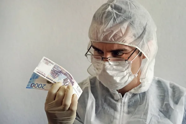 A scientist in a protective suit and medical gloves and mask holds cash in his hands. Dirt germs and virus bacteria on money.