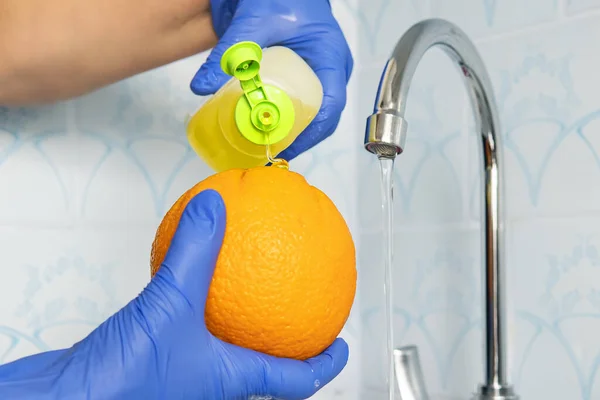 Person washes fruit with eco soap for washing in a kitchen with blue walls. Decontamination and disinfection of products from store. Prevention of coronavirus. hands in gloves wash an orange.