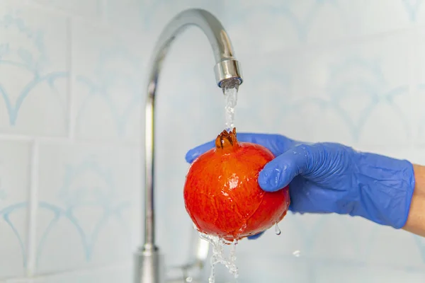 Person washes fruit with eco soap for washing in a kitchen with blue walls. Decontamination and disinfection of products from store. Prevention of coronavirus. hands in gloves wash red garnet