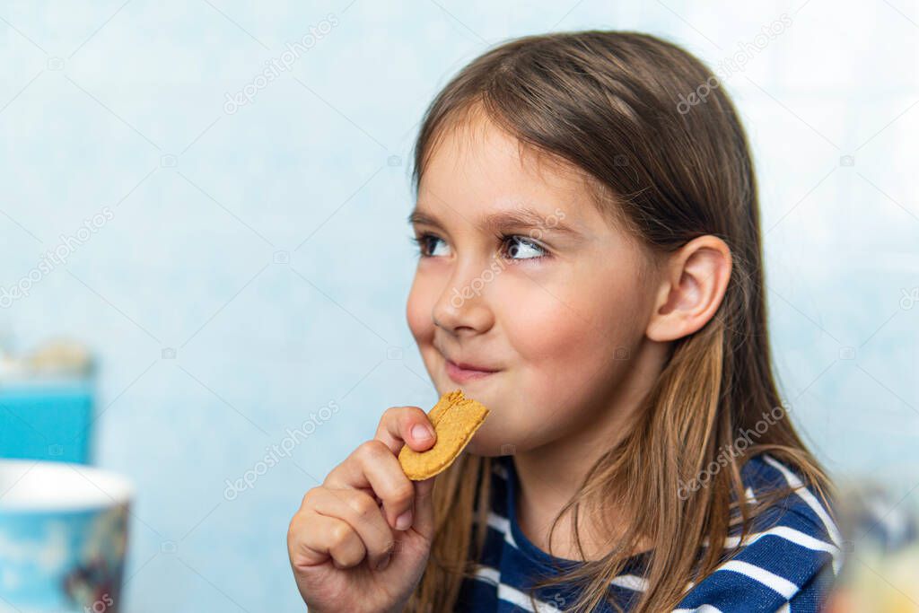 Young girl eating cookies on blue background sitting in the kitchen at home. Harmful but delicious food. Sweetness.