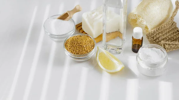 DIY ev temizliği için çevre dostu aletler ve malzemeler. Beyaz sirke, kabartma tozu, hardal tohumu, limon, saf doğal sabun, aroma yağı ve beyaz mutfak masasında luffa süngeri.