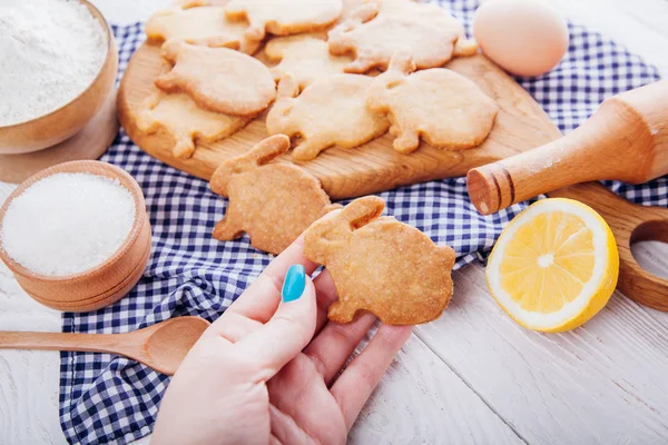 Woman holds Easter bunny-shaped cookie surrounded with ingredients.