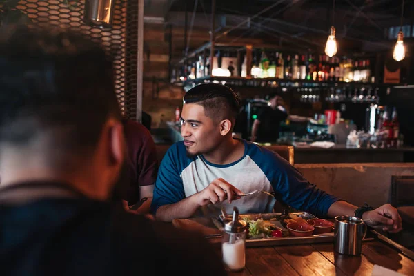 Multiracial friends eat breakfast in lounge bar. Young men chat while having tasty food and drinks.
