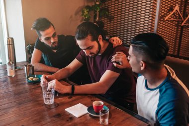 Supportive young men encourage their brokenhearted friend. Arabian guys cheer him up in restaurant. Friendship concept. clipart