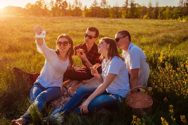 Friends eating pizza outside and taking selfie. Women and men having picnic at sunset. Fast food concept.