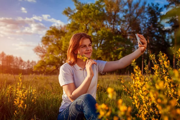 Молодая женщина делает селфи весной цветущее поле на закате. Счастливая улыбающаяся девушка расслабляется и наслаждается природой — стоковое фото