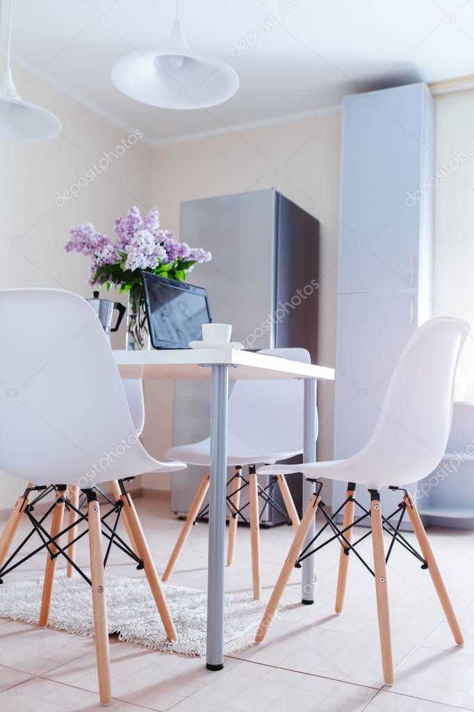 Modern kitchen design. Interior of light dining room decorated with lilac flowers. Laptop and coffee on table