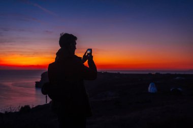 Santorini Adası 'nda gün batımında yürüyüş yapan gezgin silueti. Akıllı telefondan gece manzarasının fotoğrafını çeken bir turist.