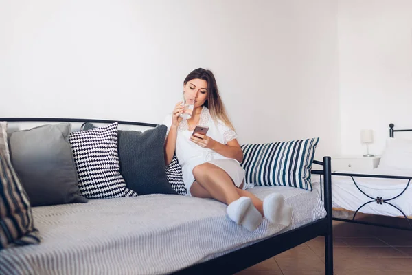 Woman relaxing in hotel living room sitting on couch using smartphone and drinking water. Morning routine