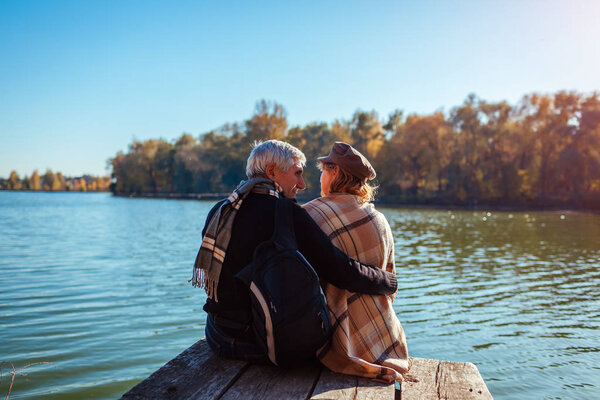 Senior family couple relaxing by autumn river. Happy man and woman enjoying nature and hugging sitting on pier
