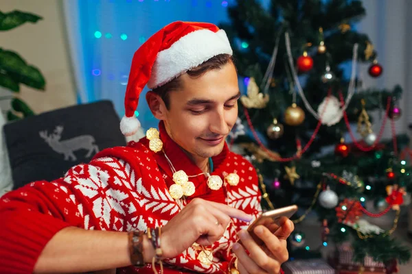 Homem de mensagens de texto amigos para o Natal usando smartphone. Um tipo a celebrar o Ano Novo sozinho em casa. Operador móvel — Fotografia de Stock