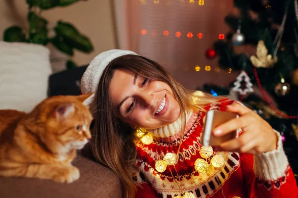 Boże Narodzenie z kotem. Kobieta robi selfie ze zwierzakiem w kapeluszu Mikołaja przez Nowy Rok drzewa w domu. — Zdjęcie stockowe