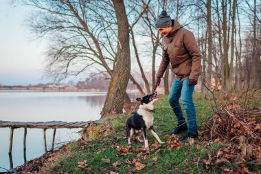 Sonbahar parkında göl kenarında köpekle oynayan adam. Mutlu hayvan dışarıda eğleniyor.