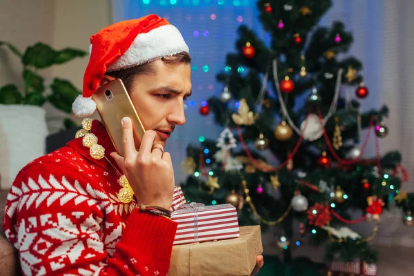 Deixado sozinho no Natal. Homem triste falando com amigos em smartphones segurando caixas de presente. Celebrando o Ano Novo em casa . — Fotografia de Stock
