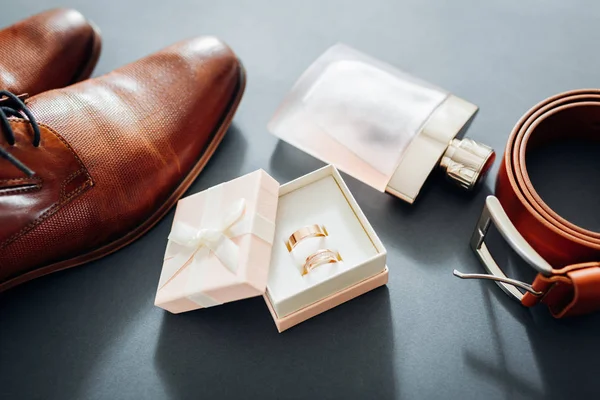 Bruidegom 's trouwdag accessoires. Bruine leren schoenen, riem, parfum, gouden ringen op grijze achtergrond. Mannelijke mode — Stockfoto