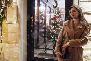 Kış tatili. Noel, yeni yıl konsepti. Şehir caddesinde aydınlatılmış bir vitrinde arkadaşlarını bekleyen bir kadın..