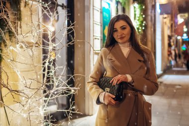 Kış tatili. Noel, yeni yıl konsepti. Şehir caddesinde aydınlatılmış bir vitrinde arkadaşlarını bekleyen bir kadın..