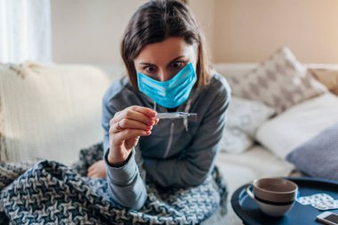 Ateşini ölçen hasta kadın. Soğuk algınlığı sırasında koruyucu maske takan bir kız, evde grip. Sağlık Hizmetleri