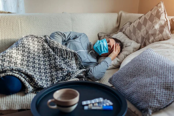 Zieke vrouw met griep of kou. Meisje liggend in bed met beschermende masker door pillen en water op tafel — Stockfoto