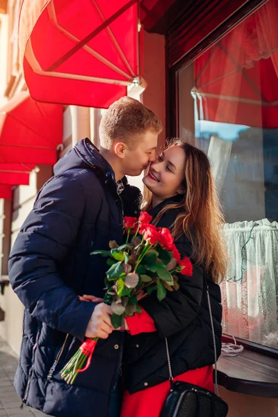 Walentynkowy pocałunek pary. Mężczyzna daje czerwone róże kwiaty dziewczynie na randkę przez kawiarnię w mieście. Dzień Kobiet — Zdjęcie stockowe