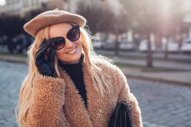 Tüylü palto giyen, güneş gözlüğü takan ve dışarıda el çantası tutan şık bir kadın. Bahar modasına uygun kadın aksesuarları..