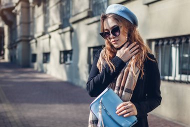 Mavi el çantası tutan şık bir kadın, bereli, güneş gözlüklü ve açık havada fular takıyor. Kadın modası. Bahar aksesuarları.