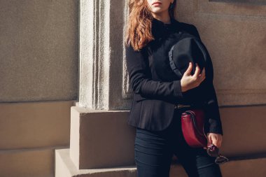 Kadın güzelliği, moda. Siyah ceket giyen şık bir kadın, duvarda kırmızı bordo çanta taşıyan bir şapka. Bahar giysileri, aksesuarlar.