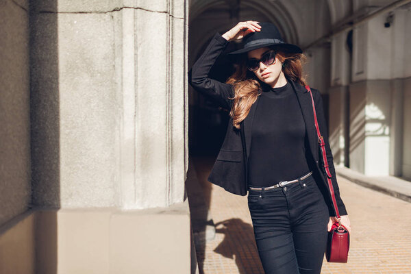 Stylish woman wearing black jacket, hat, sunglasses holding red burgundy purse handbag outdoors on street. Female beauty, fashion. Spring accessories.