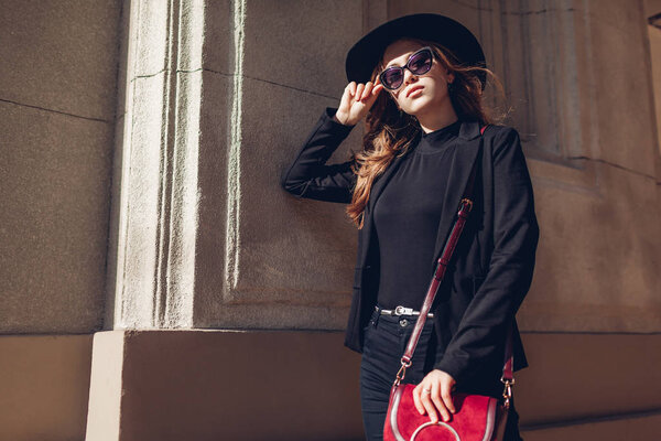 Stylish woman wearing black jacket, hat, sunglasses holding red burgundy purse handbag outdoors on street. Female beauty, fashion. Spring accessories.