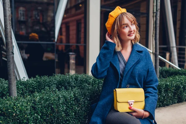 黄色のベレー帽をかぶったスタイリッシュな若い女性の肖像画は屋外で財布を保持する 現代建築による春のファッション女性のアクセサリーや服 トレンディな古典的なブルーの衣装 — ストック写真