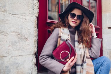 Açık havada kırmızı çantalı güneş gözlüğü tutan, şapka giyen şık genç bir kadının portresi. Bahar modası kadın aksesuarları ve bordo pencerenin yanındaki kıyafetler. Son moda klasik retro kıyafet.