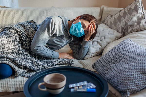 Sick woman having flu or cold. Girl lying in bed having headache wearing protective mask by pills and water on table.