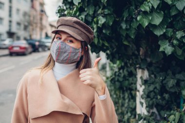 Coronavirus covid-19 salgını sırasında kadın açık havada tekrar kullanılabilir maske takıyor. Kız başparmağını boş sokakta gösteriyor. Güvende ve pozitif kalın. Bahar modası