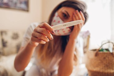 Coronavirus covid-19 salgını sırasında hamile kalmış. Koruyucu maskeli bir kadın evde hamilelik testi yaptırıyor. Şok olmuş ve üzgün bir kız. Kapat.