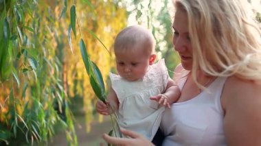Anneler Günü. Genç anne bebeğiyle birlikte bahar parkında yürüyor. Ağaç yapraklarını sallayan meraklı bebek