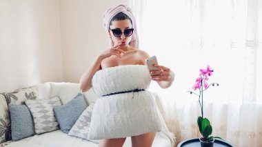 Koronavirüs salgını karantina karantinası sırasında yastık savaşı. Telefonda selfie çeken genç bir kadın yastık güneş gözlüğü takıyor ve kafasında havlu var. Kilit altında eğlenen insanlar