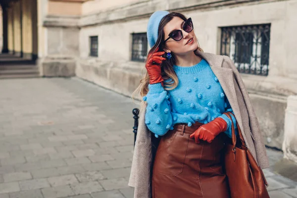 Spring vintage retro fashion female accessories and clothes. Woman wearing blue sweater beret leather skirt sunglasses holding purse on street.