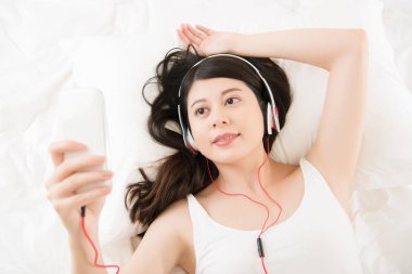 Asyalı kadın akıllı telefon kulaklık ile müzik zevk. Yatak