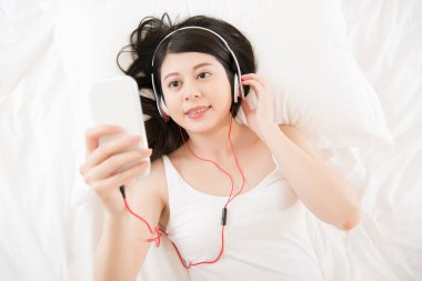 Asyalı kadın akıllı telefon kulaklık ile müzik zevk. Yatak