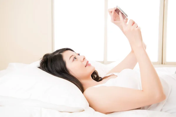 Joven sonrisa asiática mujer leyendo smart phone en cama — Foto de Stock