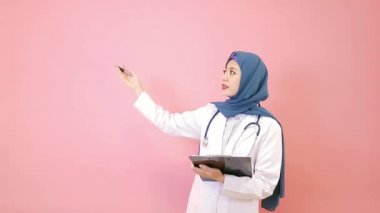 Kameraya bakarak ve telefon defteri bilgisayar ayakta pembe arka planda tutan sunu jest yapmak emin gülümseyen Müslüman doktor kadın.