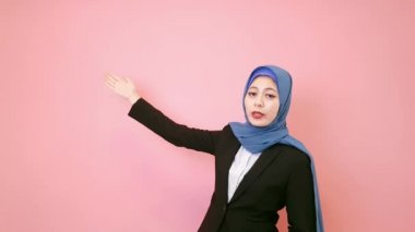 duruş ve yeni ürün açıklamak ve pembe duvar arka plan üzerinde izole kamera için yüz yapma gülümseyen genç Müslüman ofis çalışanı.