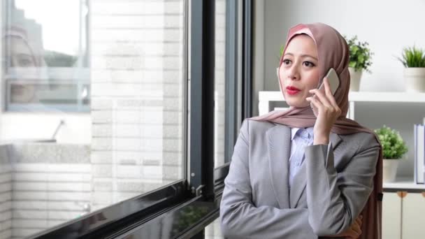 モバイルのスマート フォンを使用して若いイスラム教徒の女性ワーカーを笑って顧客説明の協力を求める場合ときウィンドウ リラックスの前に立っている彼女 — ストック動画