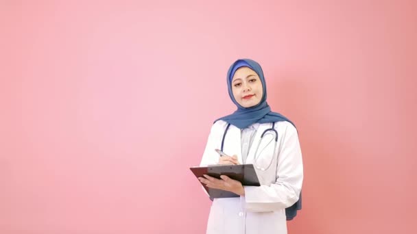 患者の医療記録の文書はピンクの背景に立って カメラ思考ソリューションを見て耳を傾ける非常に美しいイスラム教徒女医持株ペン — ストック動画