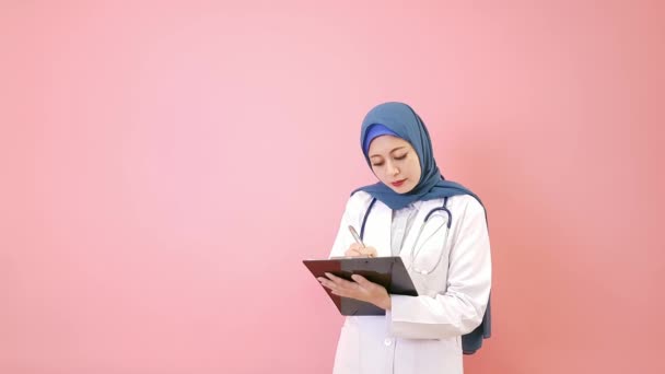 专业典雅的妇女穆斯林医生写病人病历在个人通知板隔离在粉红色的墙壁背景 — 图库视频影像