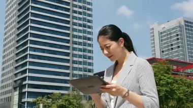 professioan Asya iş kadını yolda, arka planda Bina Finans Merkezi telefon değmek yastık dijital tablet kullanarak.