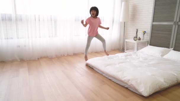 小亚洲女孩跑到床上 兴奋地跳在卧室的床上 她举起双臂享受这个赏心悦目的阳光明媚的早晨 — 图库视频影像
