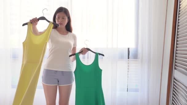 亚洲时髦的女孩犹豫买绿色礼服或黄色礼服 她站在镜子前的形象 她是如何看这两件衣服 — 图库视频影像